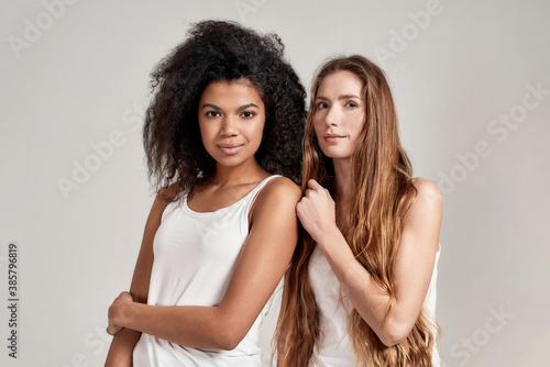 Portrait of two young diverse women wearing white shirts looking at camera while posing together isolated over grey background
