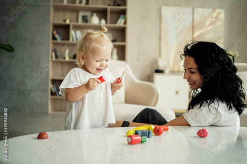 little cute blonde girl of European appearance baby and nanny brunette play with children's toys in the living room. cozy day in the apartment with the whole family.