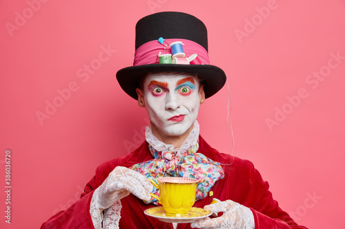 Horizontal shot of serious male hatter poses with cup of tea wears hat has manners of aristocratic gentleman dresses for masquerade carnival poses indoor. Halloween and entertainment concept