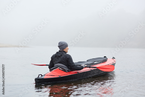 Young handsome man paddling canoe on cloudy day, canoeing on foggy day, back view of extreme sportsman enjoying water sport in autumn, looking in to distance.