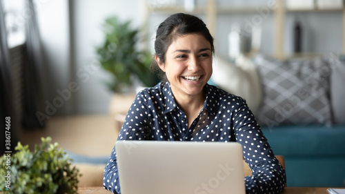 Happy cheerful millennial indian business woman sitting at desk in front of laptop laughing talking to someone in home office room looking into distance feels overjoyed. Successful freelancer portrait