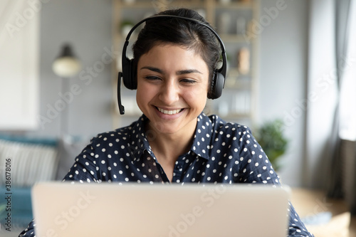 Cheerful Indian woman e tutor sit at desk home office wear headset looks at laptop talk by video call, helpline employee and pleasant chat to client remotely, distance webinar online teaching concept