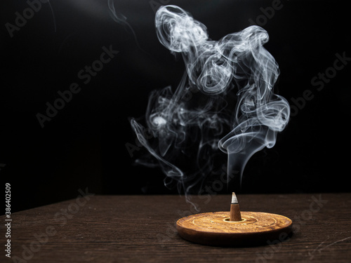 Burning cone incense on wooden holder on dark surface