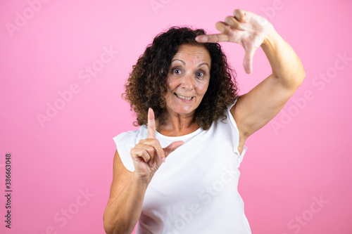 Middle age woman wearing casual white shirt standing over isolated pink background smiling making frame with hands and fingers with happy face