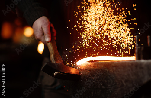 The blacksmith hits the red-hot workpiece in the forge with a hammer and glowing sparks fly in all directions