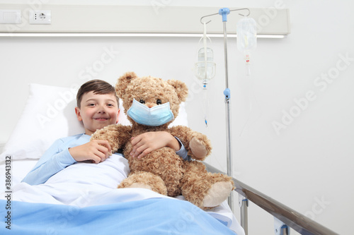 Smiling child in hospital bed with teddy bear wearing protective mask, corona virus covid 19 protection concept 