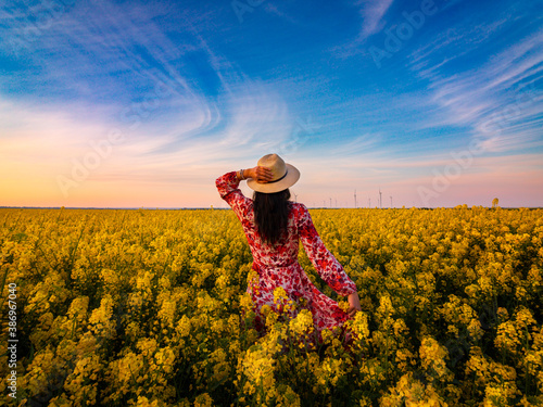 Back view young women with long hair and hat on head standing with open arms in field full of yellow flowers