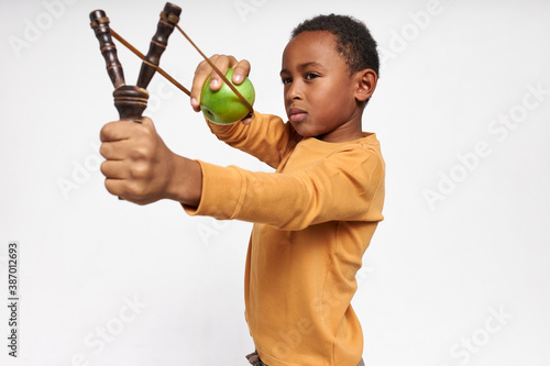 Isolated image of serious concentrated little black boy holding Y-shaped stick with elastic, shooting green apple, having focused facial expression. Accurate African child playing with slingshot
