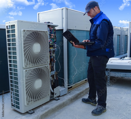 air conditioning technician making a diagnosis of an industrial air conditioning unit with a laptop next to other VRV condenser units on a rooftop in a sunny day