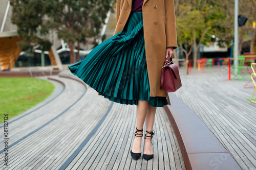 Fashionable young woman wearing green pleated midi skirt, sweater, high heel shoes, beige wool coat and holding burgundy handbag in hand on the city street. Street style.