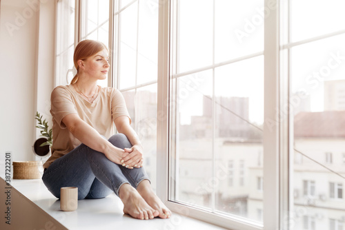 Woman is sitting on the windowsill daydreaming with cup of coffee and looking to the beautiful view of the city