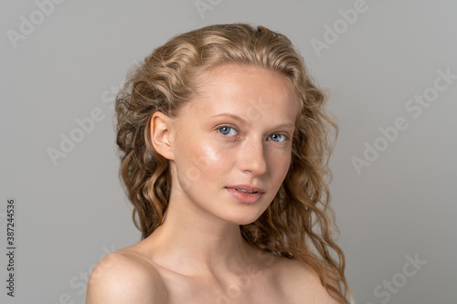 Closeup of pretty young woman face with blue eyes, curly natural blonde hair, has no makeup, touching her soft skin, standing shirtless with bare shoulders, looking at camera. Studio grey background