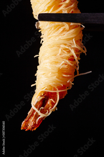 Delicious appetizer shrimp fried in noodles held by metal kitchen tongs on a black background
