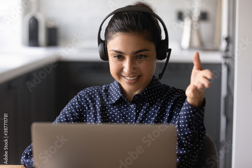 Smiling Indian woman in earphones work on laptop consult client customer online from home office. Happy ethnic female in headphones talk speak on video call on computer. Web communication concept.