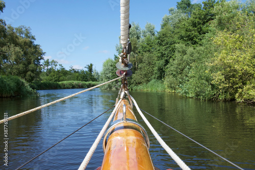 The bowsprit of a sailing yacht on a sunny tree lined river