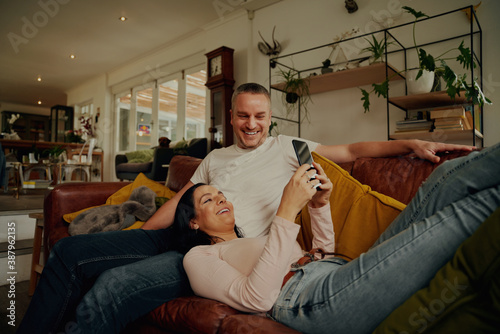 Cheerful young wife using phone while lying on husband's lap and relaxing on couch at home