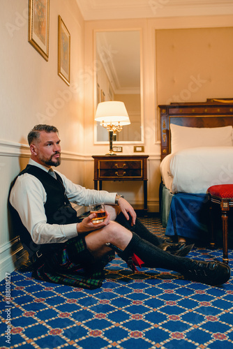 handsome mature courageous stylish man (gay) scotsman in kilt drinking whiskey on the floor in fancy hotel room. Style, fashion, lifestyle, culture, travel, ethnic concept.