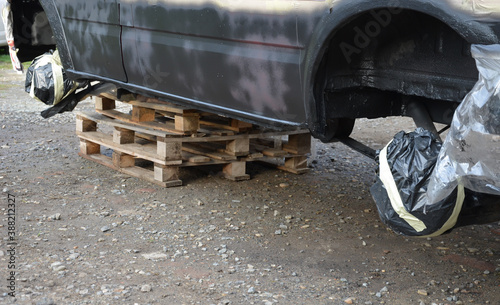 repair of a red white car and spraying black chassis against rust. the car is sealed with tape and foil. the car painter also disarmed the car wheels. The car is built on wooden pallets. stolen wheels