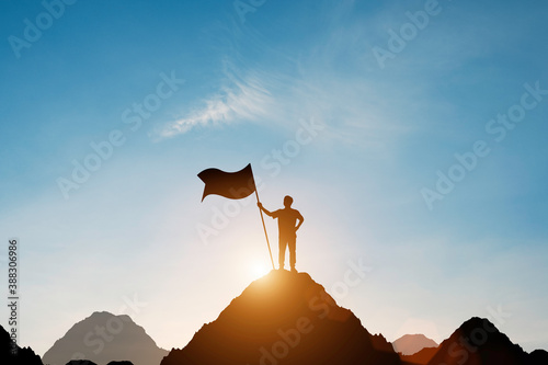 Silhouette of businessman holding flag on the top of mountain with over blue sky and sunlight. It is symbol of leadership successful achievement with goal and objective target.