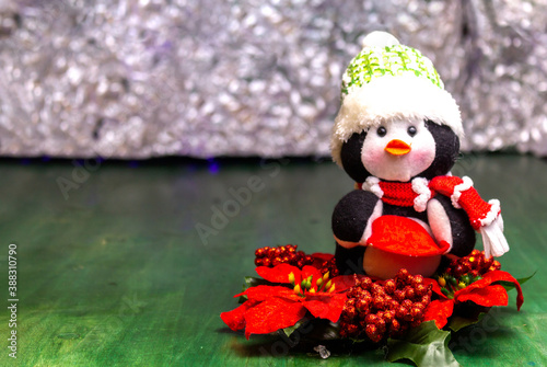 Christmas doll, penguin with wool hat on a Christmas flower on a green wooden table on a luminous background