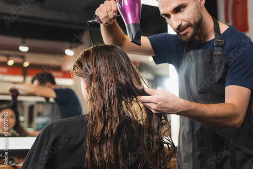 young bearded hairstylist drying hair of woman near blurred mirror reflection