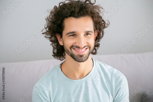 Joyful handsome curly haired guy wearing casual t-shirt, sitting on couch at home, looking away, smiling and laughing. Male portrait concept