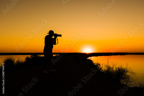 silhouette of a photographer with reflex camera and large lens in a lake taking pictures with the sunset in the background