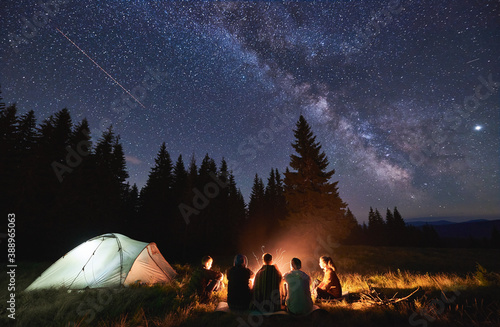 Evening summer camping, spruce forest on background, sky with falling stars and milky way. Group of five friends sitting together around campfire in mountains, enjoying fresh air near illuminated tent
