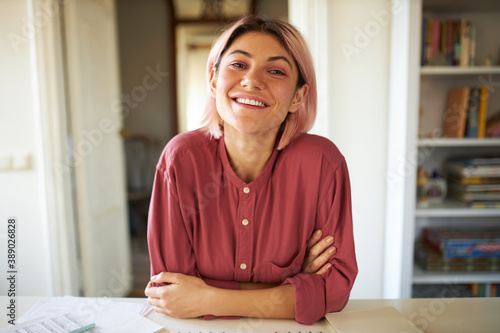 Positive cheerful young woman posing in cozy home interior, sitting at table with papers, working distantly, looking at camera with broad smile, having online group meeting with colleagues