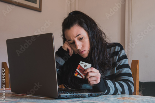 Una mujer joven usa un ordenador portátil para realizar compras por internet mientras sostiene una tarjeta de crédito con una expresión de confusión