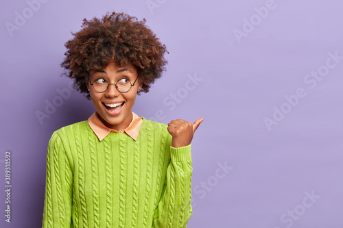 Happy good looking ethnic woman with Afro hair points away on blank space shows place for your advertising content or commercial text dressed in casual jumper isolated over purple backround.