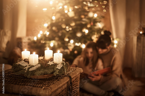 Child sits with mother in front of the Christmas tree and read a book together and look forward to Christmas, advent wreath