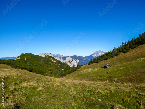 A panoramic view on the valley in Kaiserau Kreuzkogel region, Austrian Alps. There is a small cottage on the side of a meadow. Few trees on the slopes, endless mountain chains. Sunny and bright day.