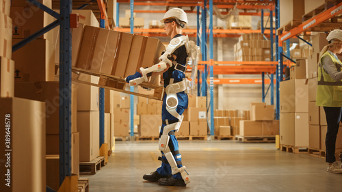 High-Tech Futuristic Warehouse: Worker Wearing Advanced Full Body Powered exoskeleton, Lifts Heavy Pallet full of Cardboard Boxes. Delivery Exosuit amplifies strength.