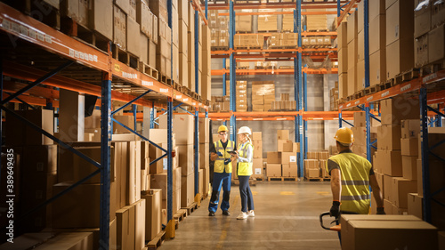 Retail Warehouse full of Shelves with Goods in Cardboard Boxes, Male and Female Managers Use Digital Tablet, Talk about Product Delivery. In Background Forklift Working in Logistics Storehouse