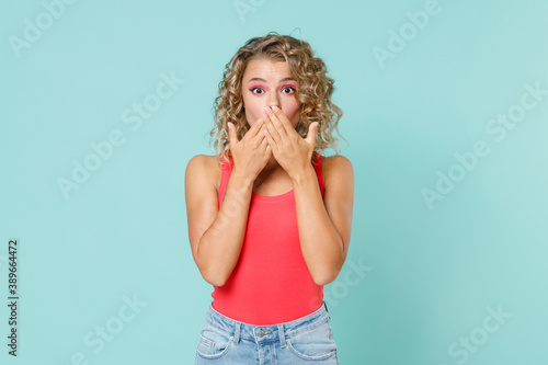 Shocked amazed beautiful young blonde woman 20s wearing pink basic casual tank top standing covering mouth with hands looking camera isolated on blue turquoise colour wall background studio portrait.