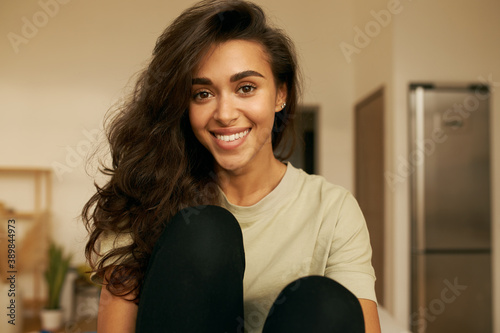 Indoor shot of beautiful happy young 20 year old Arabic woman relaxing at home wearing comfy clothing, looking at camera with broad smile, showing white straight teeth. Cute girl having rest indoors