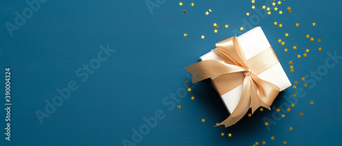 White gift box with golden ribbon bow on blue background with confetti. Christmas present, valentine day surprise, birthday concept. Flat lay, top view.