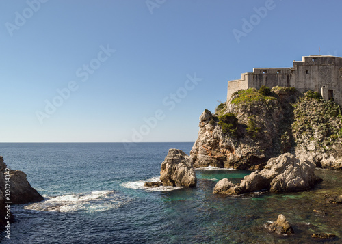The sea, some rocks and a part of the historic city wall in Dubrovnik, Croatia. The sky is blue.