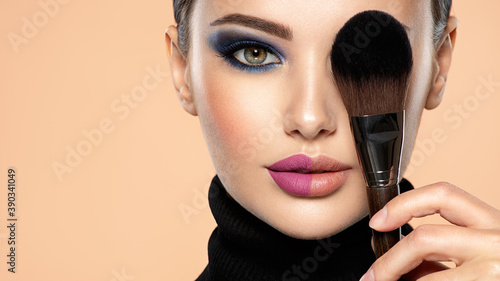 Portrait of a girl with cosmetic brush at face. Woman covering one eye on the face using makeup brush. One half face of a beautiful white woman with bright makeup and the other is natural.