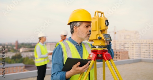 Caucasian man topographer in casque measuring angle with total station on building roof. Male builder constructor doing topographic measures and using tablet device. Geodesic concept. Device computer.