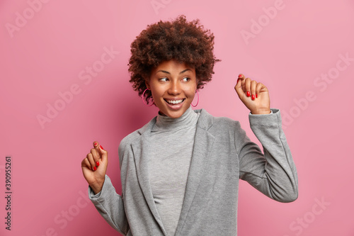 Indoor shot of successful female winner dressed in grey formal clothing makes triumph dance smiles broadly looks aside poses against pink background. Executive woman worker rejoices success.