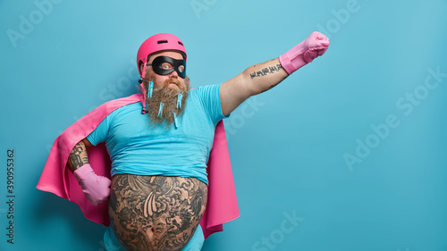 Self confident serious bearded man ready to help people wears superhero costume pretends being heroic character has supernatural power stretches arm over blue background with empty blank space