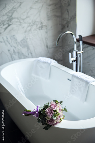 A delicate wedding bouquet of roses lies on the edge of a snow-white bathroom. Designer bathroom in the center of the bathroom. White artificial stone bathtub. Designer bathroom interior
