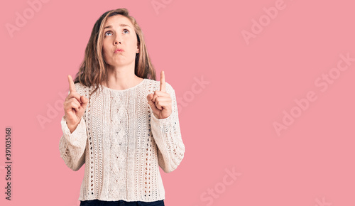 Young beautiful blonde woman wearing casual sweater pointing up looking sad and upset, indicating direction with fingers, unhappy and depressed.