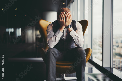 A man entrepreneur is in sorrow because of his dismissal; a businessman on an orange armchair near an office window covered his face with his hands after learning about the bankruptcy of the company