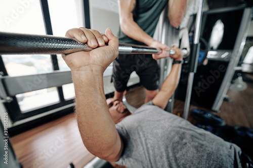 Close-up image of strong man lifting heavy barbell, his trainer standing near by