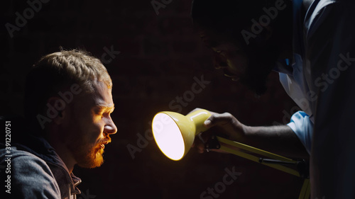 Police officer pressing and interrogating man suspected of crime glowing light of lamp into face.