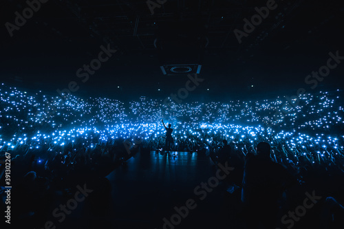 Vocalist in front of crowd on scene in stadium. Bright stage lighting, crowded dance floor. Phone lights at concert. Band blue silhouette crowd. People with cell phone lights.