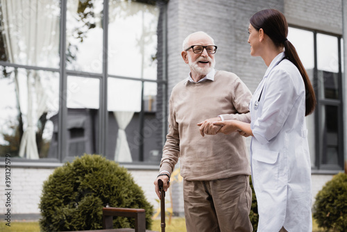 geriatric nurse supporting aged man strolling with walking stick